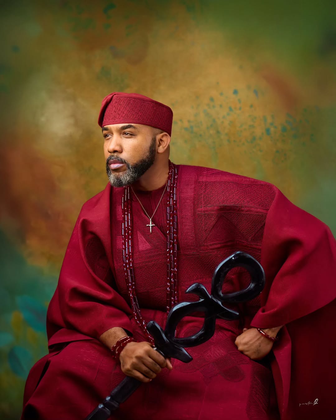 Close-up portrait of Banky W in a deep crimson agbada and fila holding a ceremonial Yoruba staff.