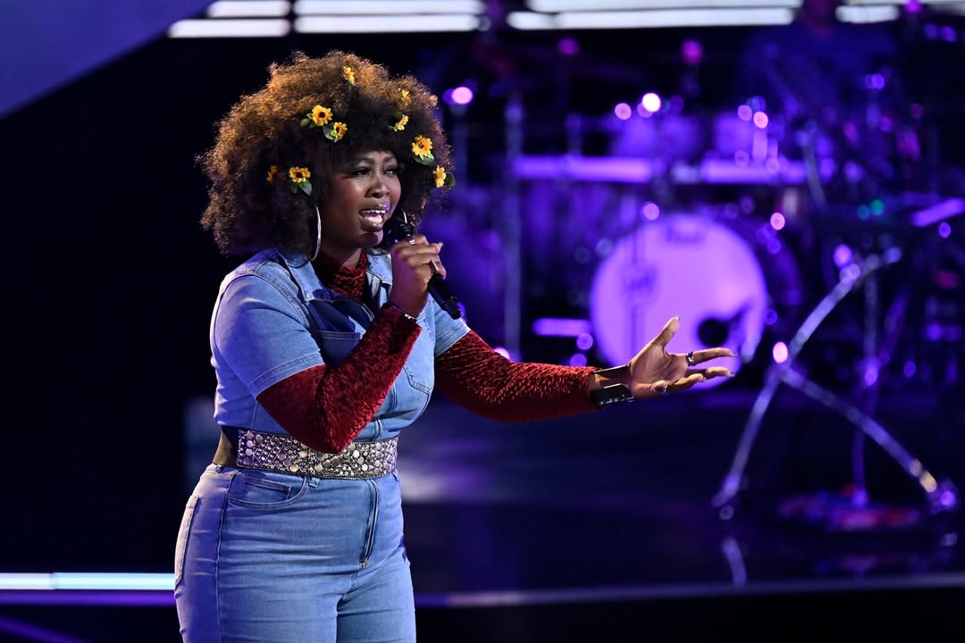 Alexia Jayy performing on The Voice stage in a denim jumpsuit with red textured sleeves and sunflowers in her hair.
