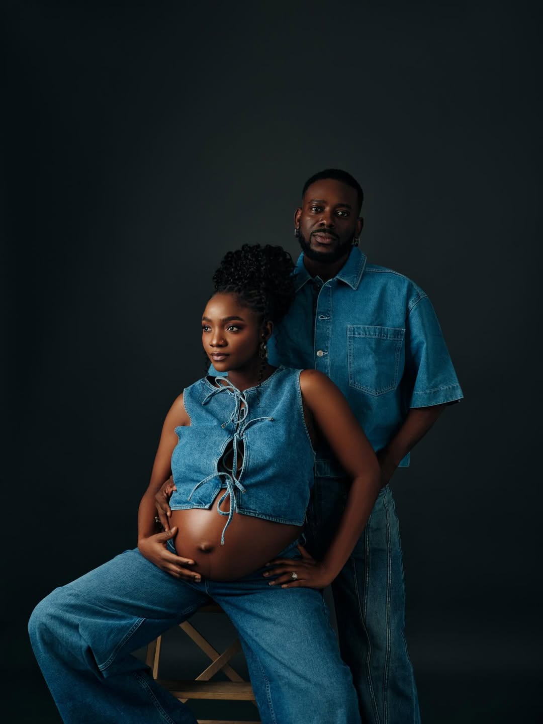 Simi and Adekunle Gold in matching denim outfits. Simi is seated, wearing a denim crop top and wide-leg jeans, showing her baby bump. Adekunle stands beside her in a denim shirt and jeans.