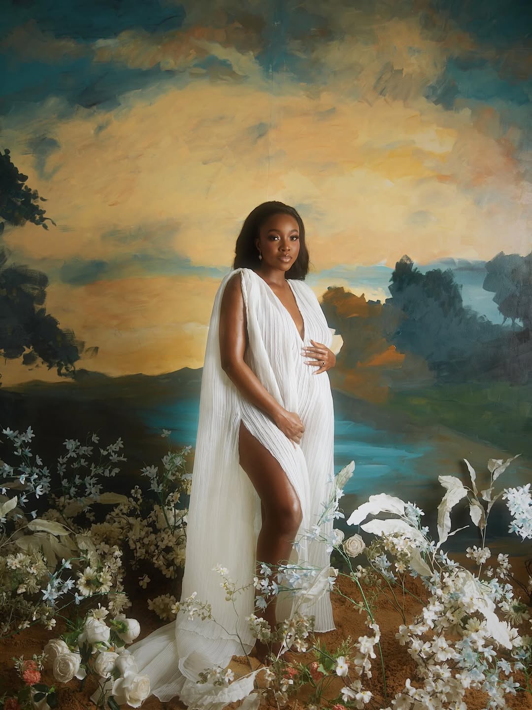 Actress Ini Dima-Okojie in a flowing white pleated Toju Foyeh gown, posing against a painted landscape with white roses for a maternity photoshoot.
